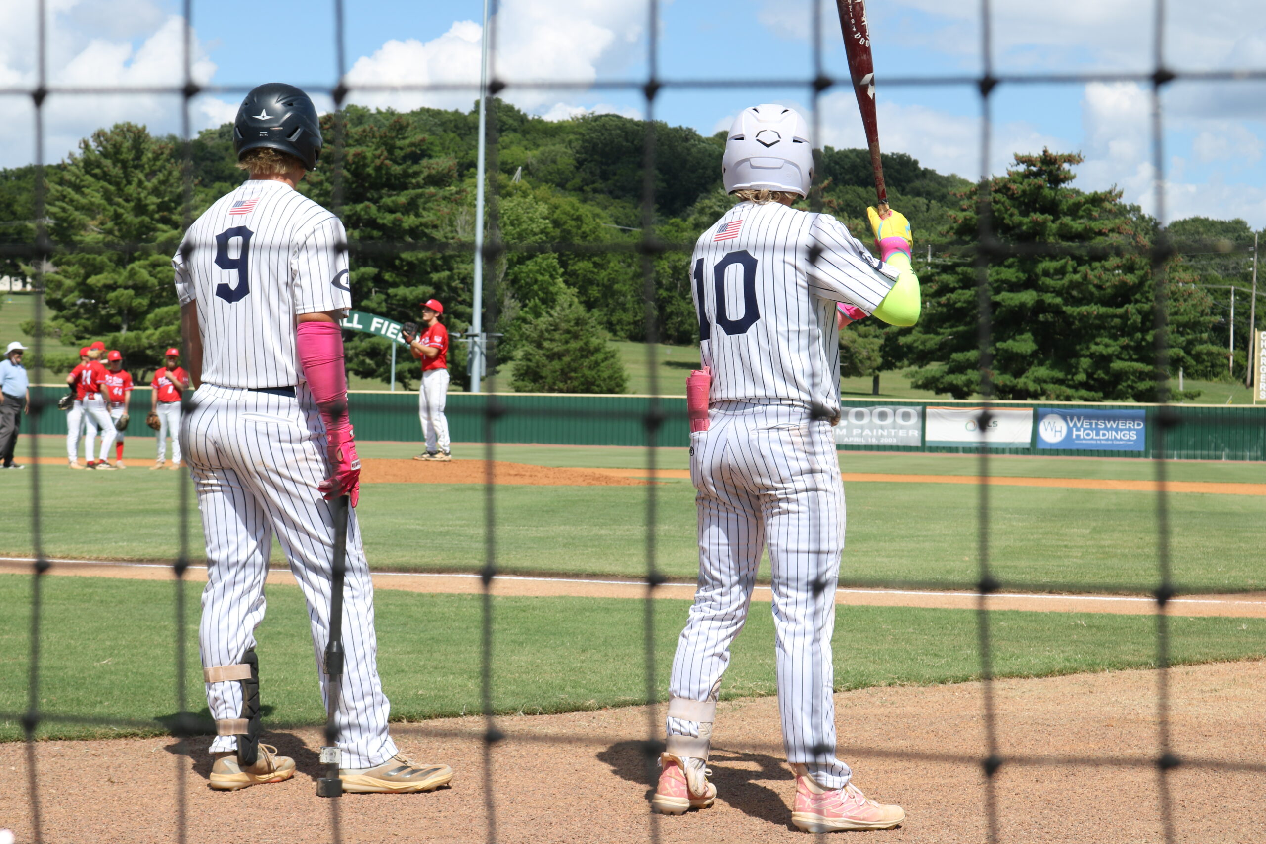 Two baseball players in striped uniforms stand at the on-deck circle, one holding a bat. The scene captures an afternoon game with teammates in the background.