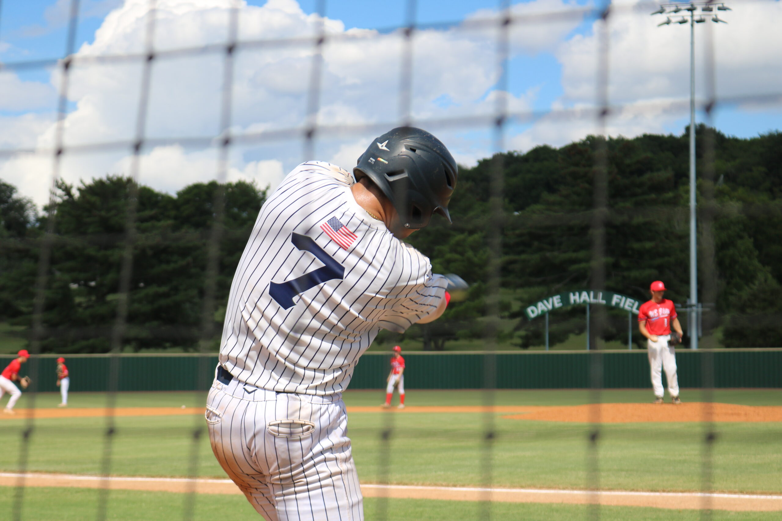 A baseball player in a striped jersey and helmet swings at a pitch, with a pitcher in red uniform on the mound at Dave Hall Field.