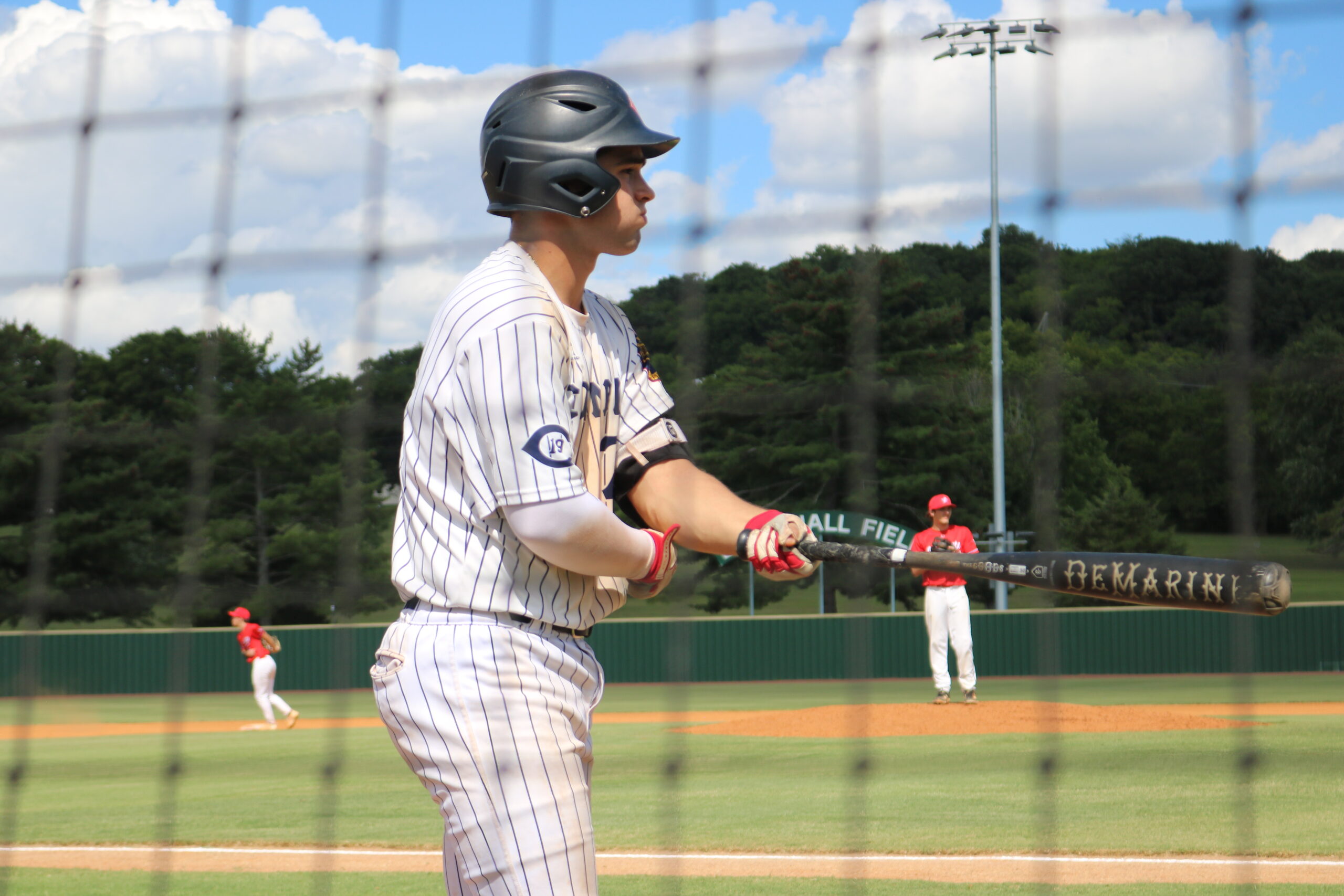 A baseball player in a pinstriped uniform prepares to bat, while a red-uniformed pitcher stands in the background on a sunny day.